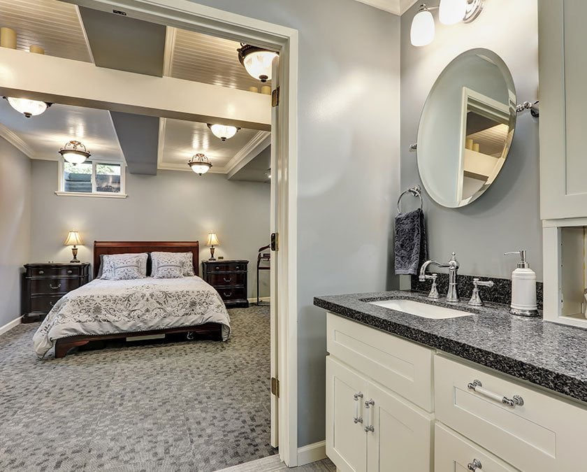 Basement bedroom with carpet floor, large bed, modern lighting, bathroom with white cabinets, quartz countertop, and round mirror.