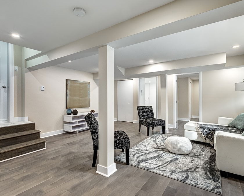 Basement family room with wooden floor, light beige painted walls, two gray chairs, white and black carpet, and white couch.